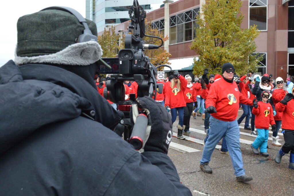 Holiday parade camera operator in foreground looking towards people walking wearing red shirts