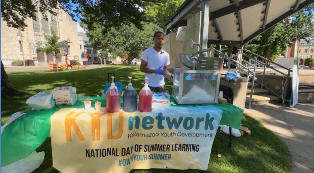 Black male at ice cone table with National Day of Summer Learning banner in Bronson Park