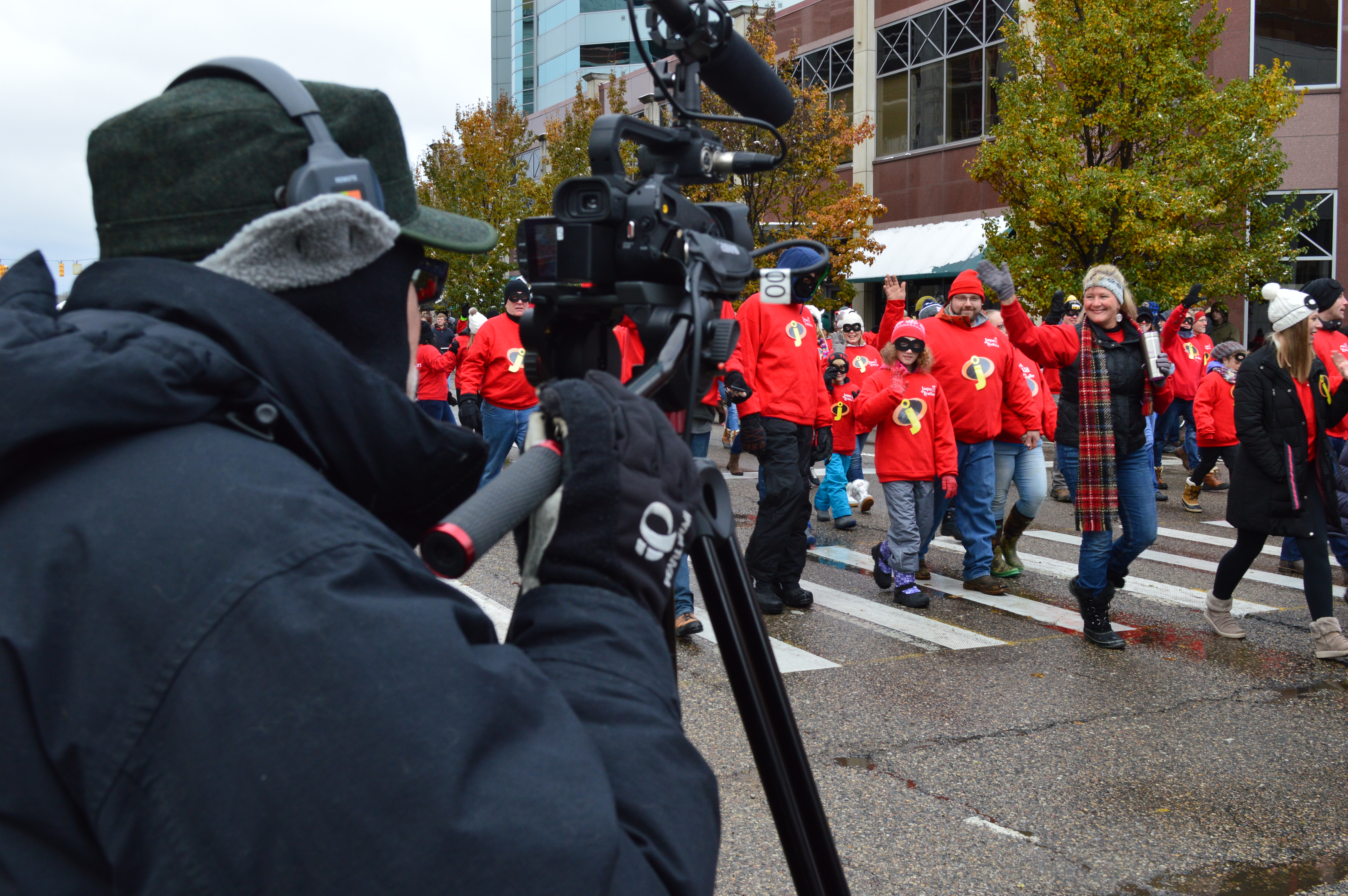 Camera operator in foreground with parade walkers wearing red in background and person waving