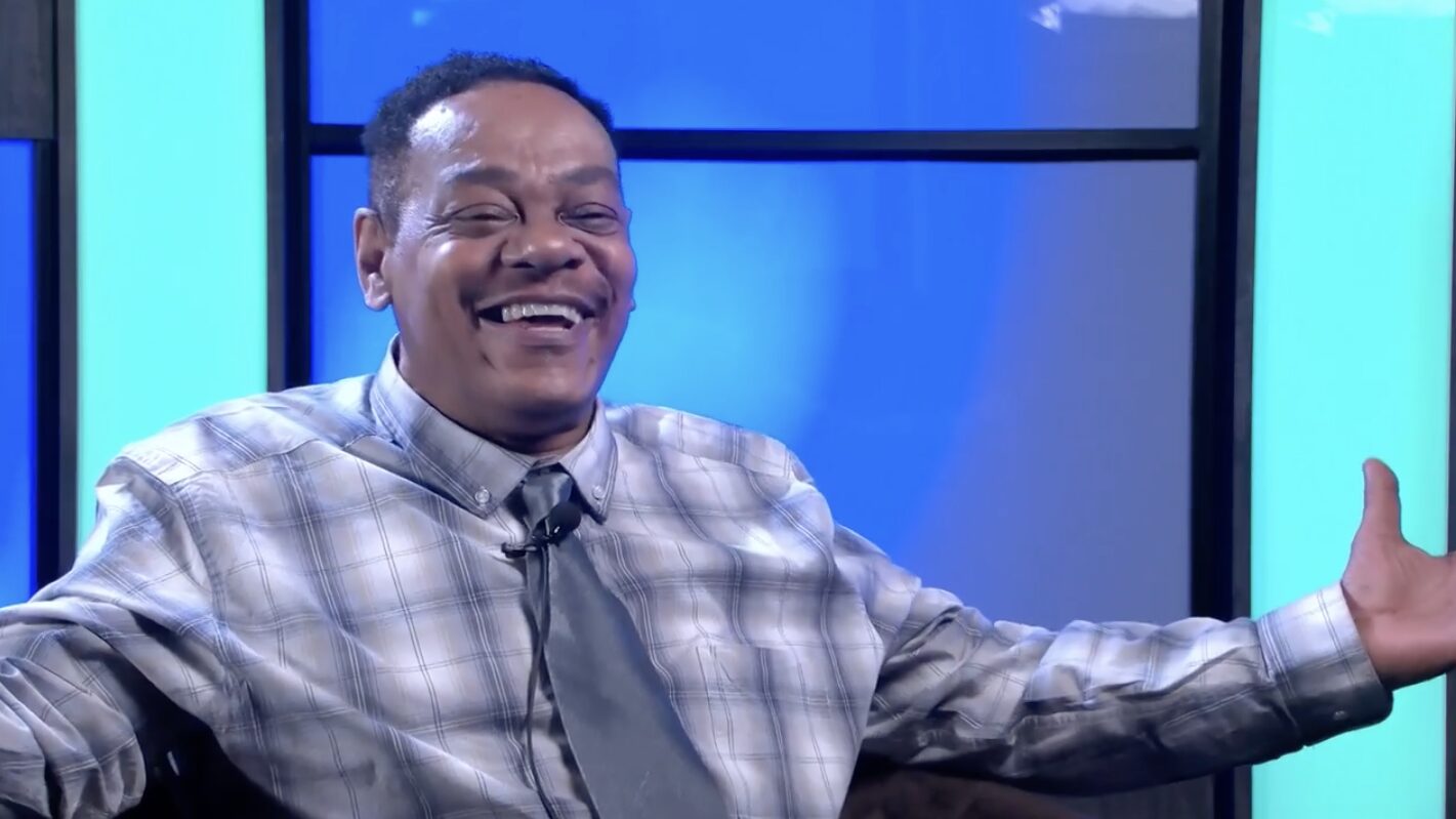 A photo of a smiling man wearing a tie, with his arms outstretched in a welcoming gesture. He is sitting in the TV studio at PMN.