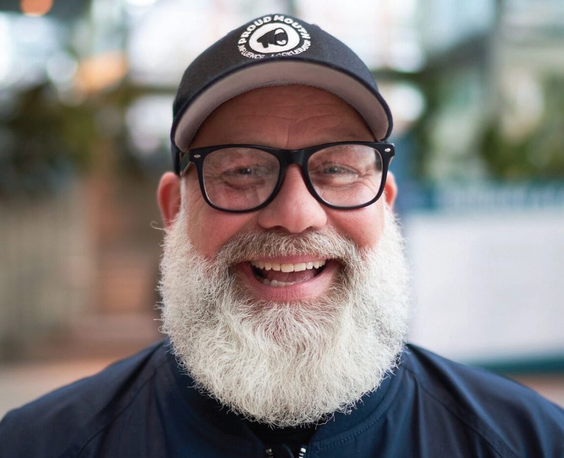 Matt, a man wearing a ball cap, black glasses, and a black tshirt, sporting a big white beard, smiles at the camera.