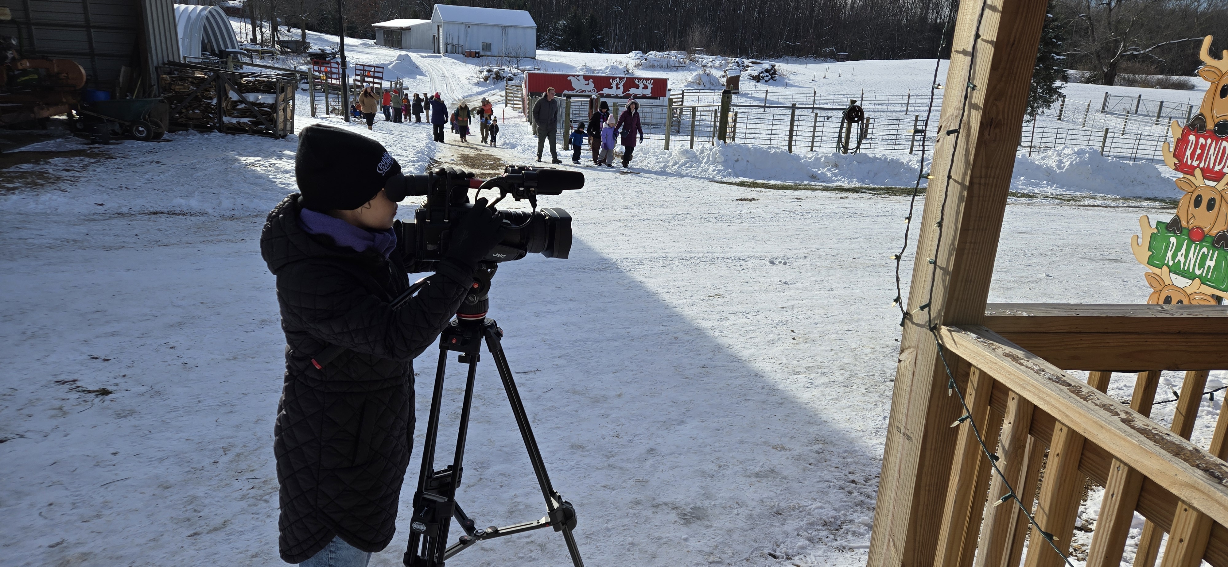 A person wrapped up in a coat and hat, face looking into a big newscamera which is sitting on a tripod. There is snow on the ground, and people are seen milling around in the background, as well as fences, suggesting a farm.