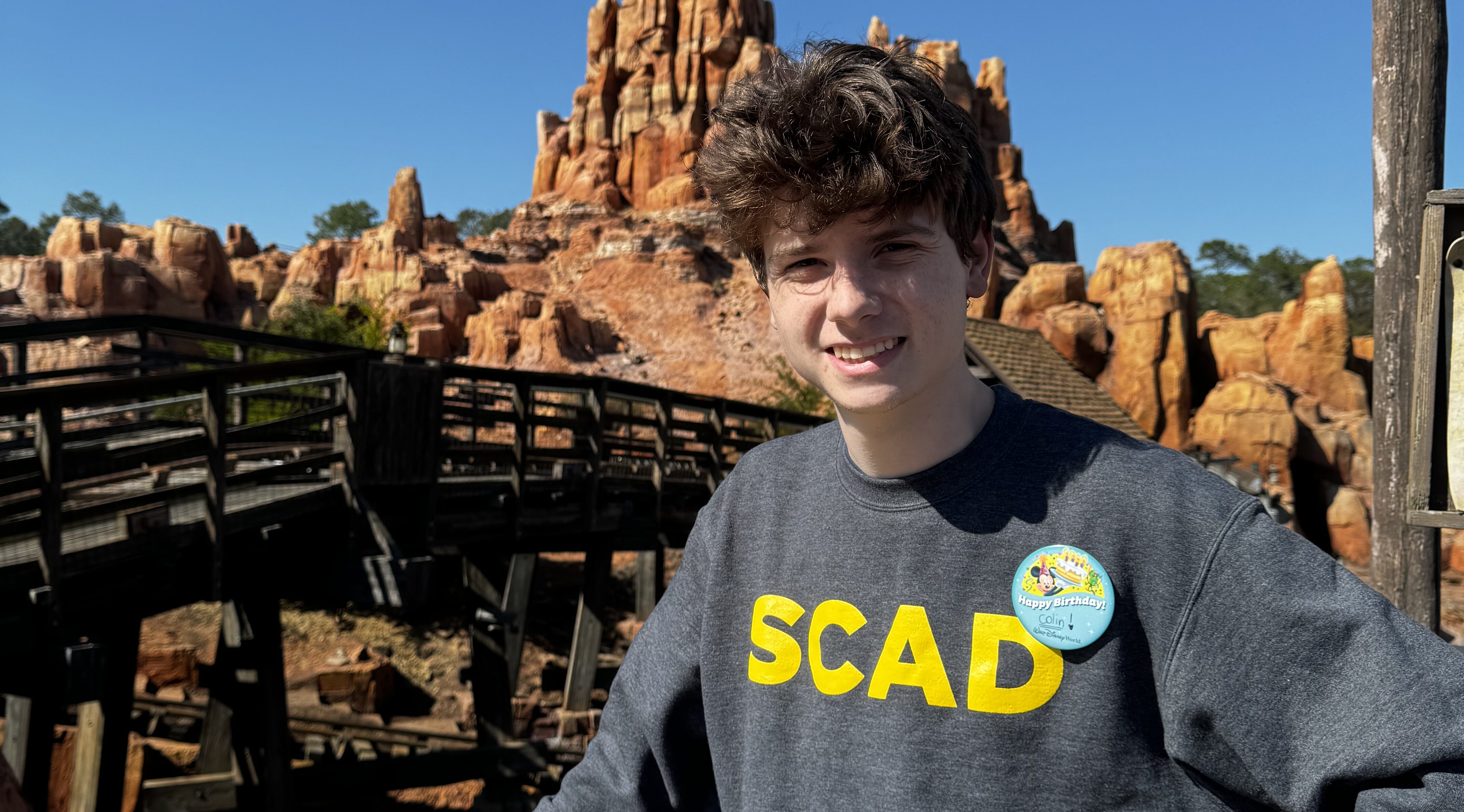 Colin, wearing a "SCAD" sweatshirt standing in a theme park that looks like a rocky desert.