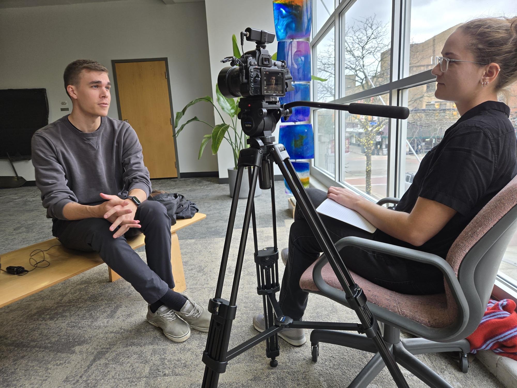 A young man sits on a bench on the left side of the photo, with a camera pointed towards him, while he talks to a young woman on the other side of the camera sitting in a chair and holding a notebook on her lap.