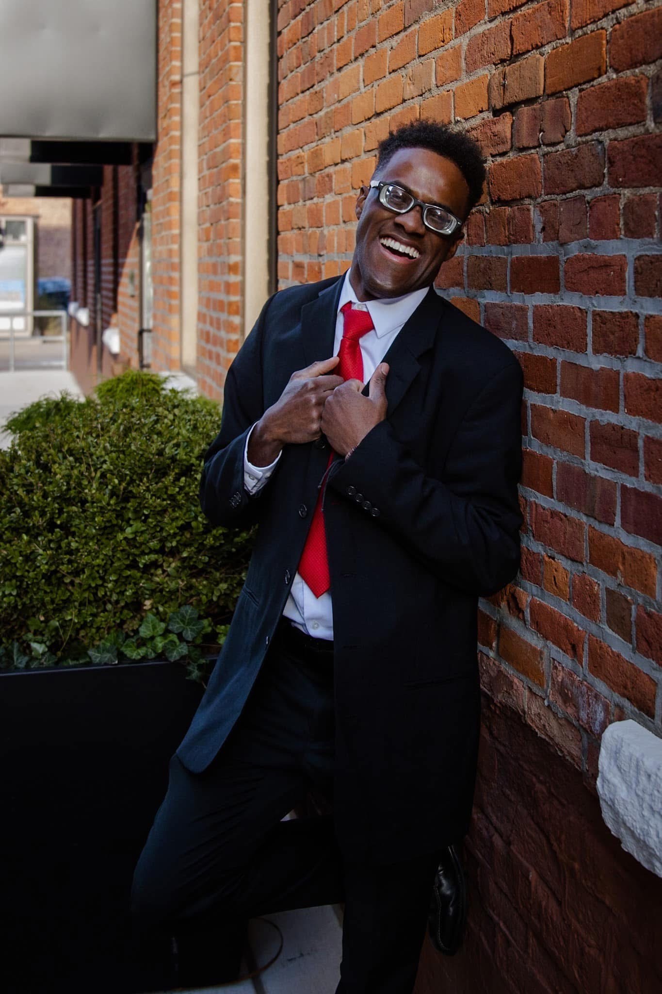 Tyrell, a young black man wearing glasses and a suit and tie, holds the lapels of his jacket while leaning against a brick wall, his face mid-laugh.