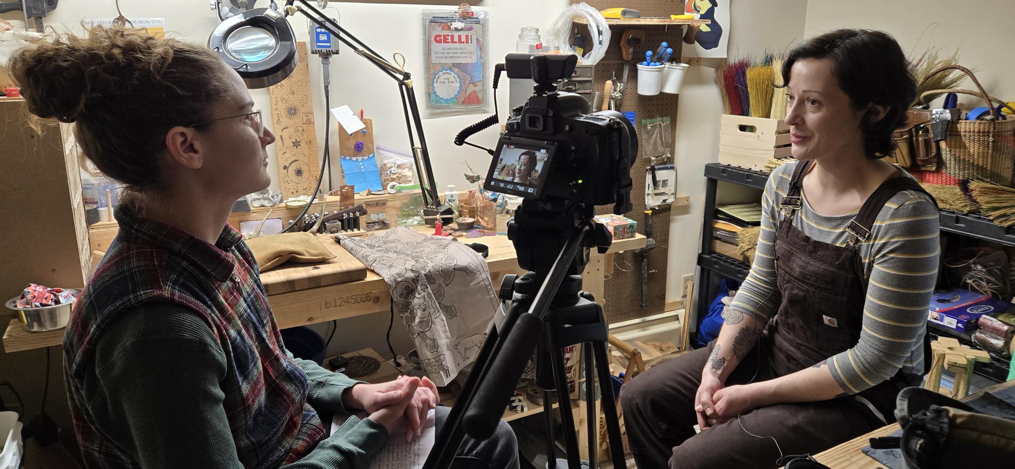 A young woman with blond hair tied into a bun, wearing glasses and a plaid shirt, interviews and points a camera at another woman with dark hair wearing overalls. They are seated in a workshop, with lots of scattered tools and materials on a work bench and the wall behind them.