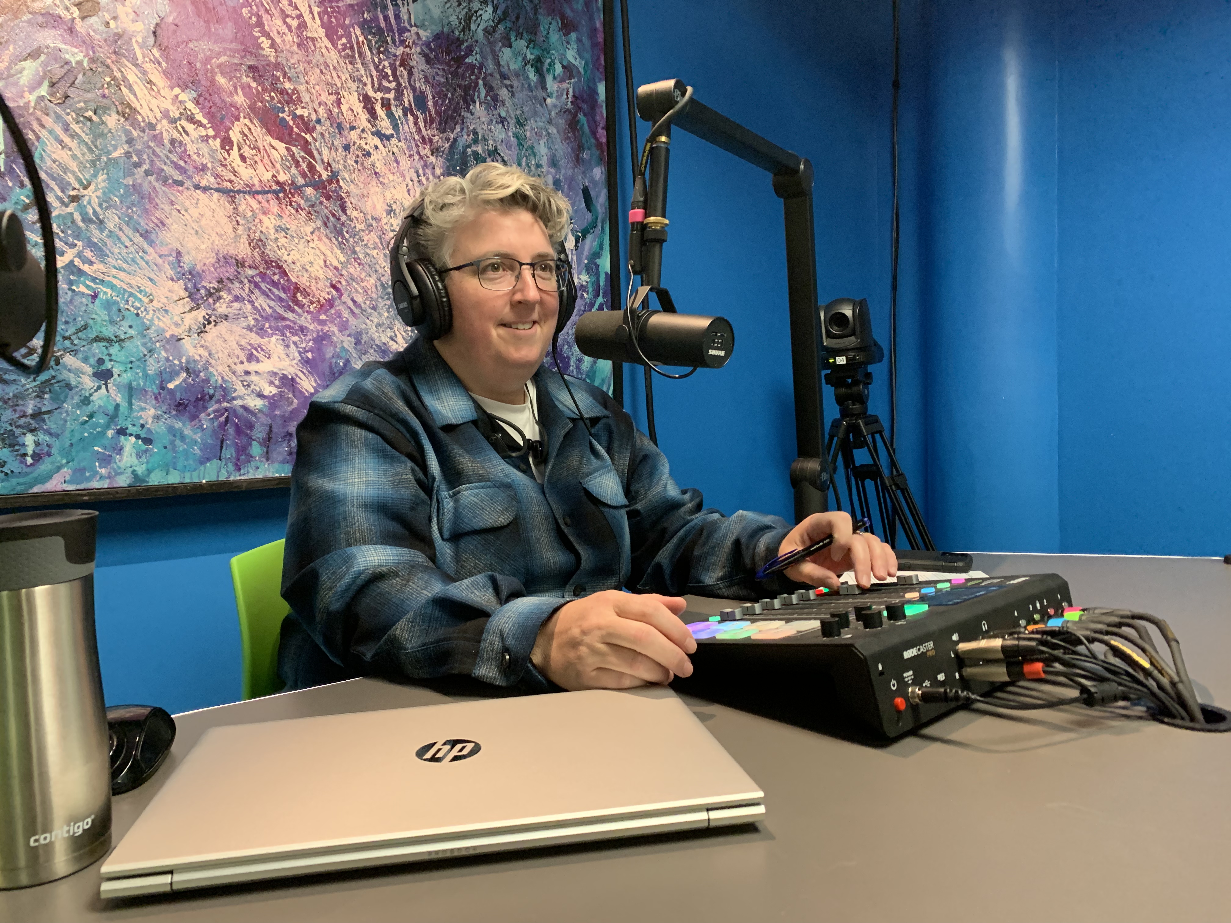 A photo of Kristen smiling, while sitting in the Vidcast Studio, wearing headphones and sitting in front of a microphone.