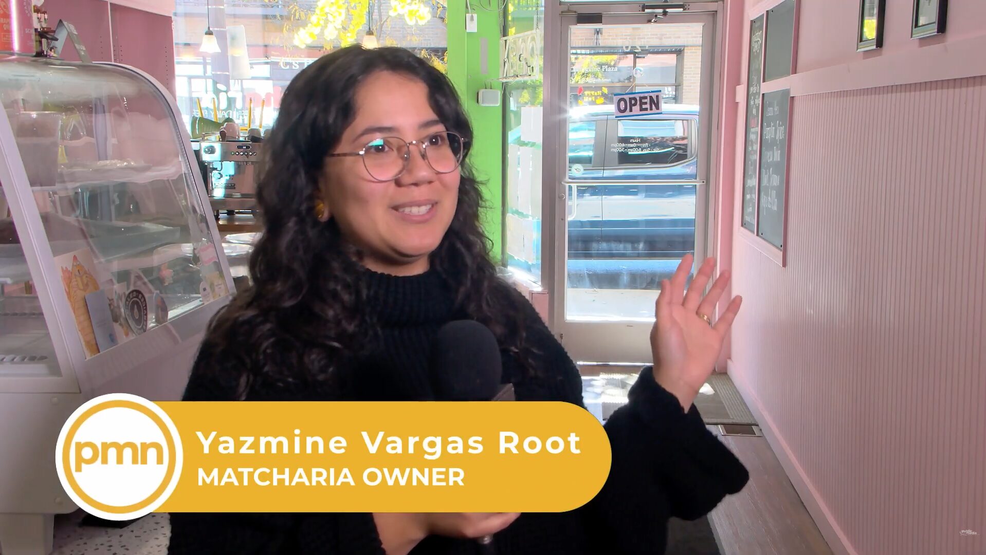 A young woman with glasses holding a microphone smiles. Behind her is a coffee shop with pink walls. A lower-third at the bottom reads "PMN" and "Yazmine Vargas Root, Matcharia Owner"