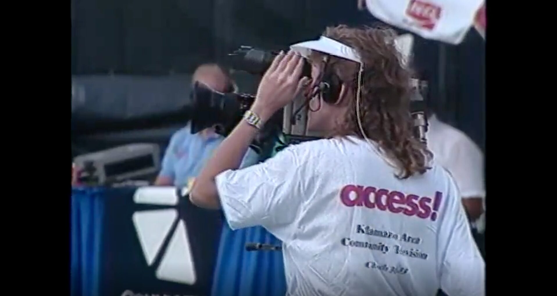 A person with long curly hair, wearing a visor, a watch, a headset, and a white t-shirt that reads ''access! Kalamazoo Area Community Television'' on the back. They are looking into the viewfinder of a big camera, their hand up to touch the camera. The quality of the picture is low, indicating it's an older photo.