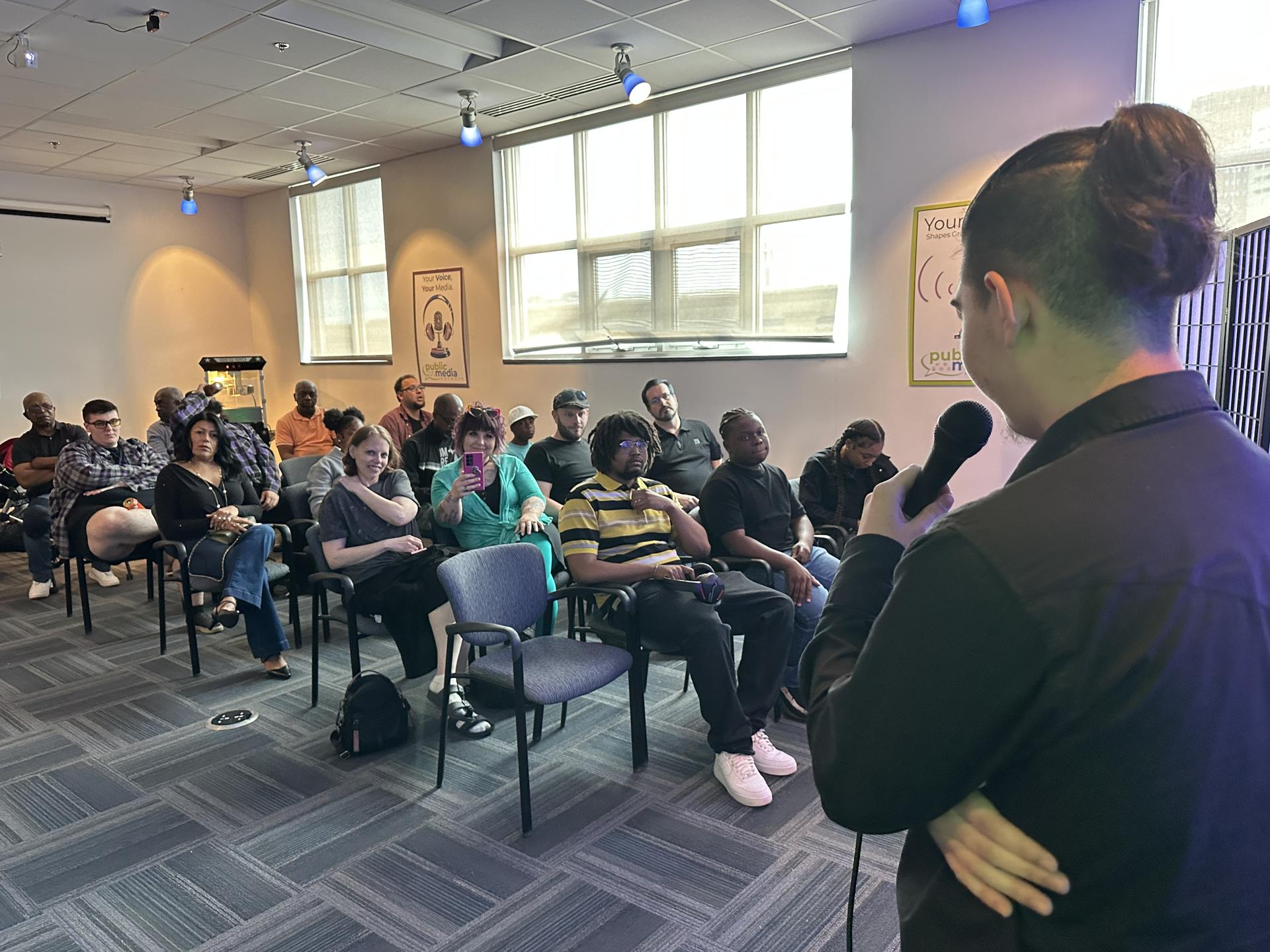 A room full of seated people look towards a young person who stands holding a mic.