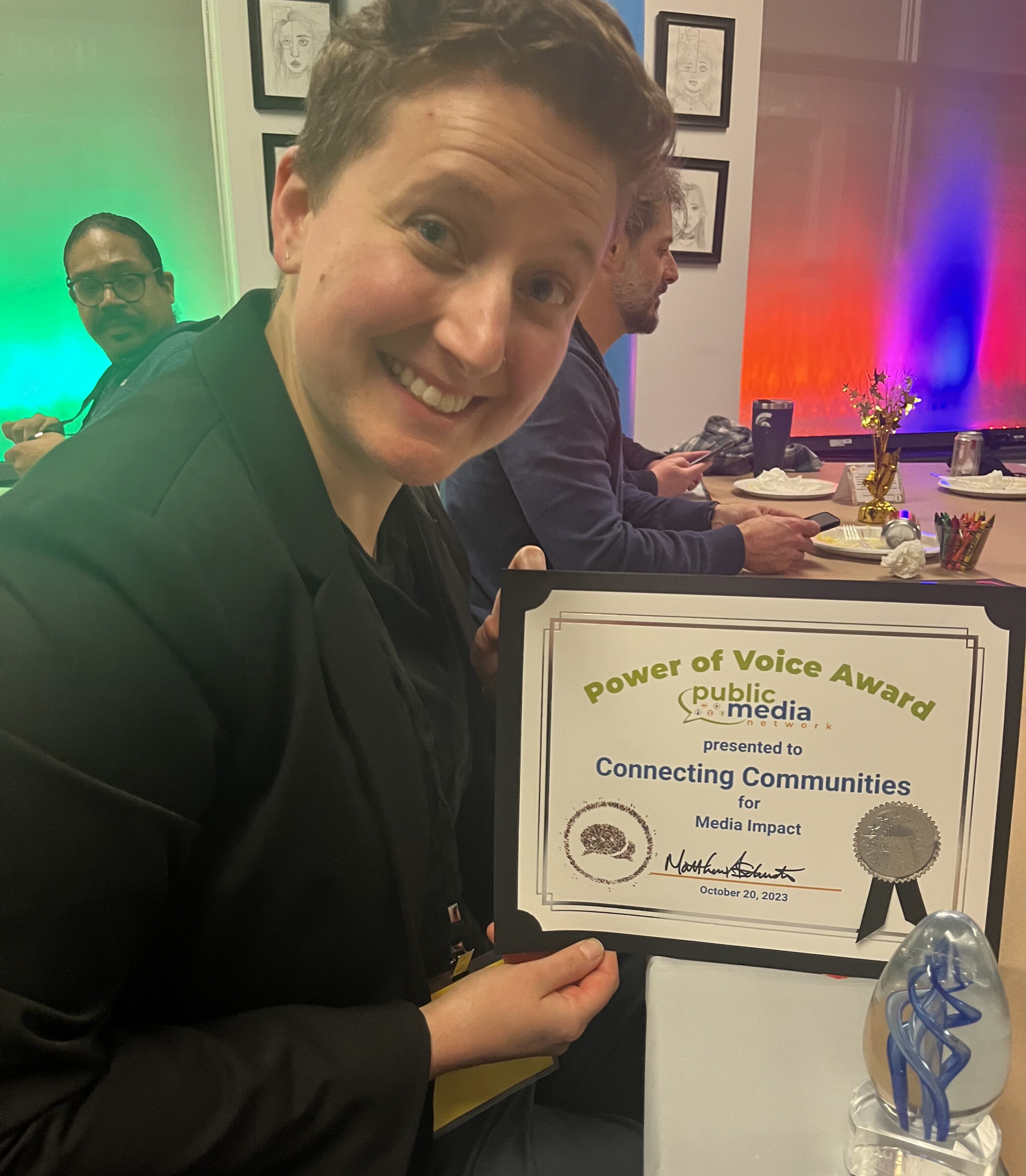 Dee smiles, holding a plaque that reads 'Power of Voice Award presented to Connecting Communities for Media Impact' and 'Public Media Network'