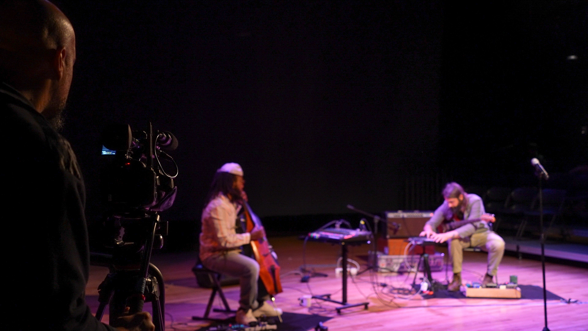 A man operates a camera which is pointed towards a lit up stage where two people holding instruments sit in chairs.