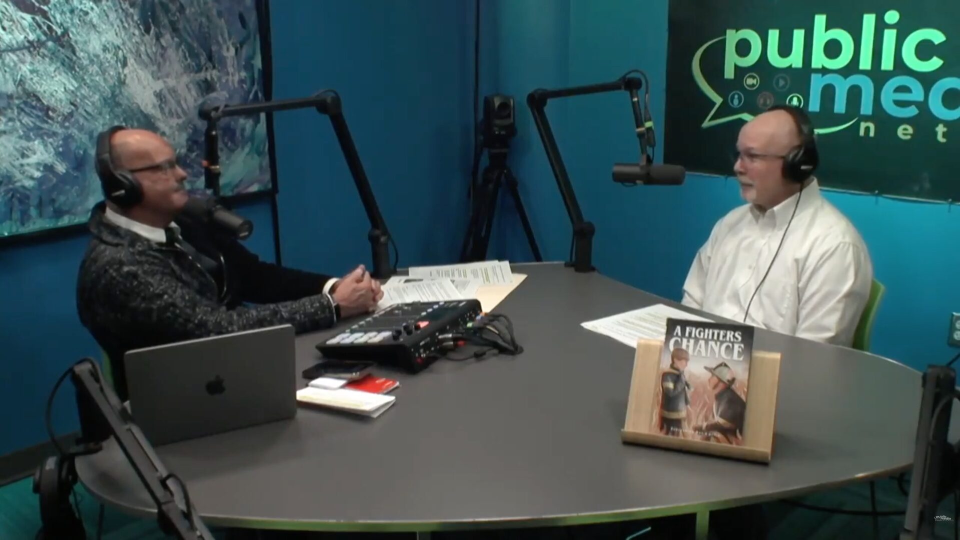 Two men sit at the table in the podcast studio, looking at each other. Both are wearing headphones and speaking into microphones. The man on the left has a laptop in front of him, and the man on the right has a book sitting near him titled "A Fighter's Chance".