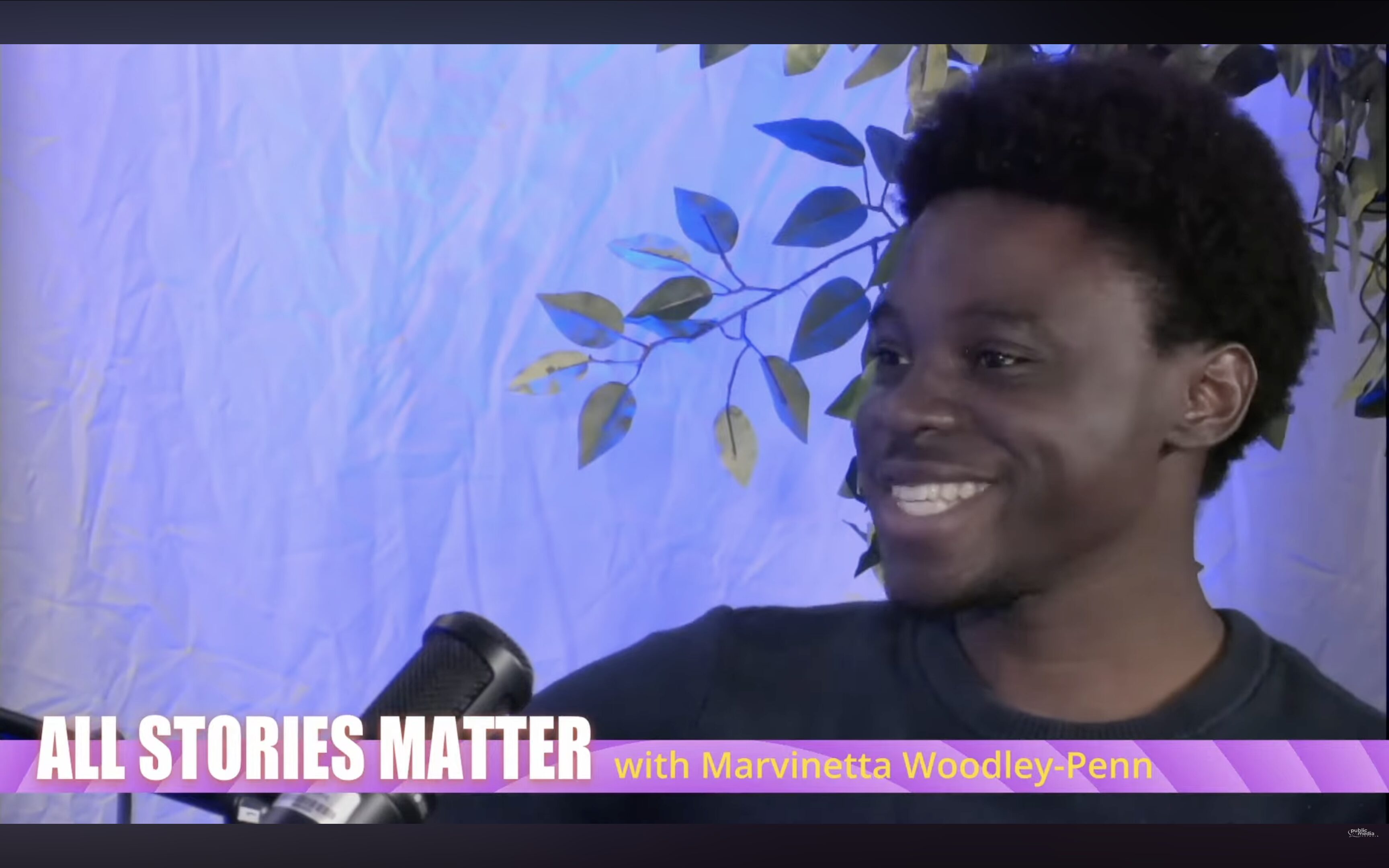David F. Gakodi gives a big smile. He is a young Black man wearing a black t-shirt with a microphone in front of him. A pink banner at the bottom of the photo reads ALL STORIES MATTER with Marvinetta Woodley-Penn